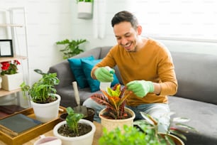 Good-looking man using a spray bottle to water his plants while sitting on his sofa living room. Hispanic man enjoying his gardening hobby at home