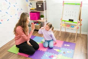 Female tutor giving a high five to a little girl while sitting on the foam mat of the playroom. Caucasian teacher tutoring an elementary girl