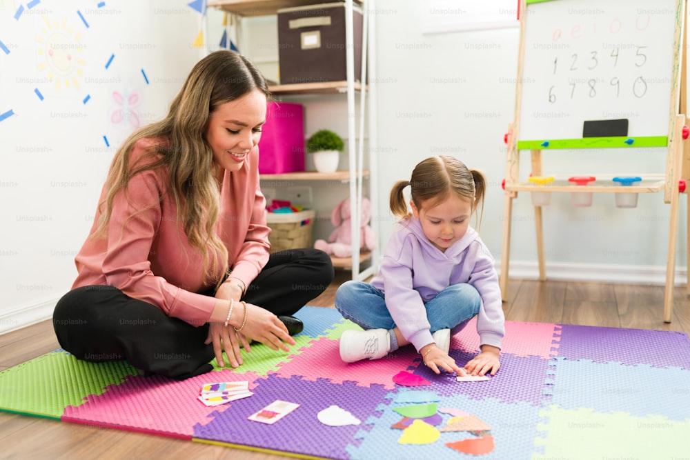 백인 여성 심리학자가 초등학생과 함께 바닥에 앉아 아동 치료 세션 동안 시퀀스 퍼즐을 푸는 것을 돕고 있다