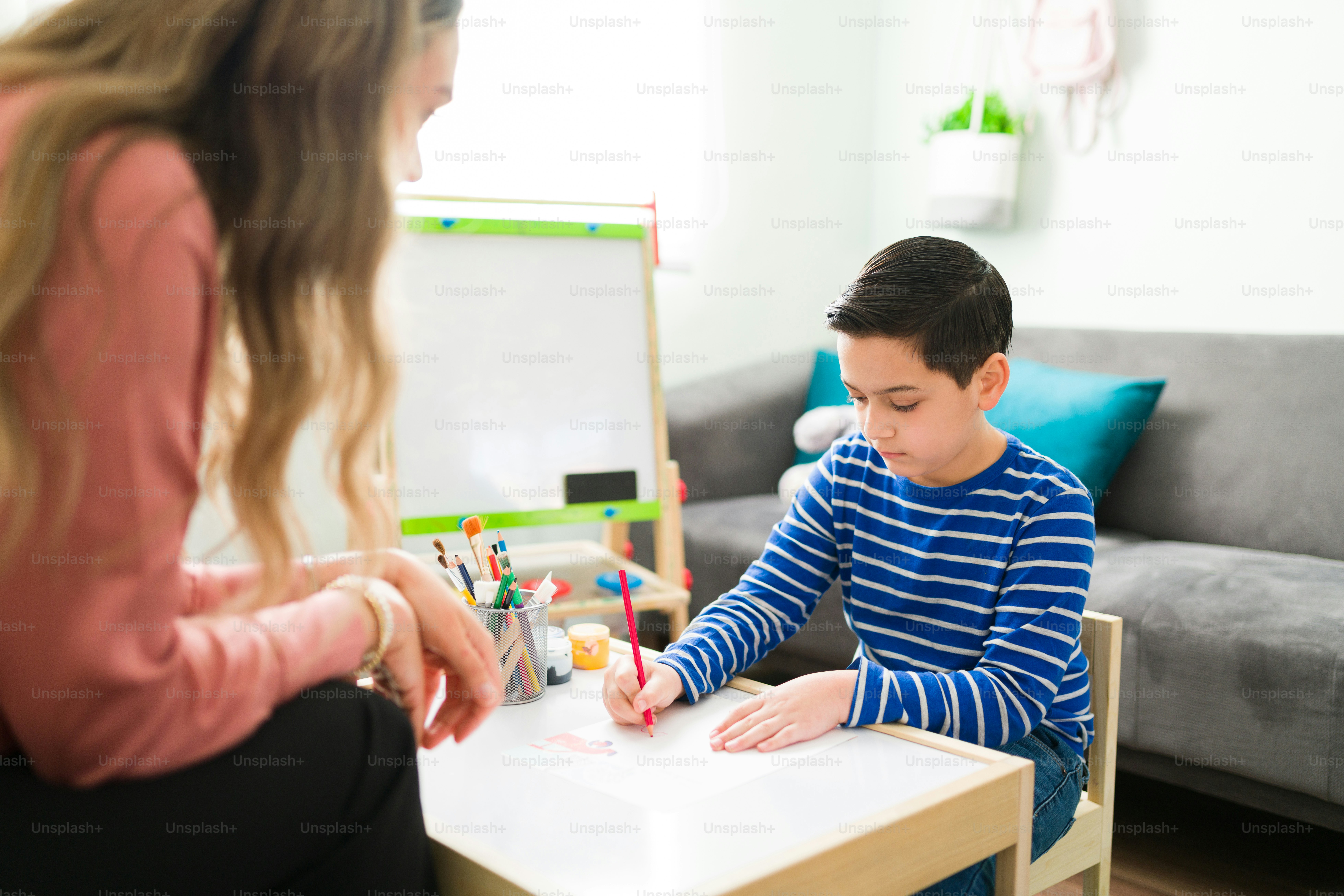 Handsome little boy working on a drawing while sitting at a small table with his female therapist. Sad boy during an art session at his psychologist office