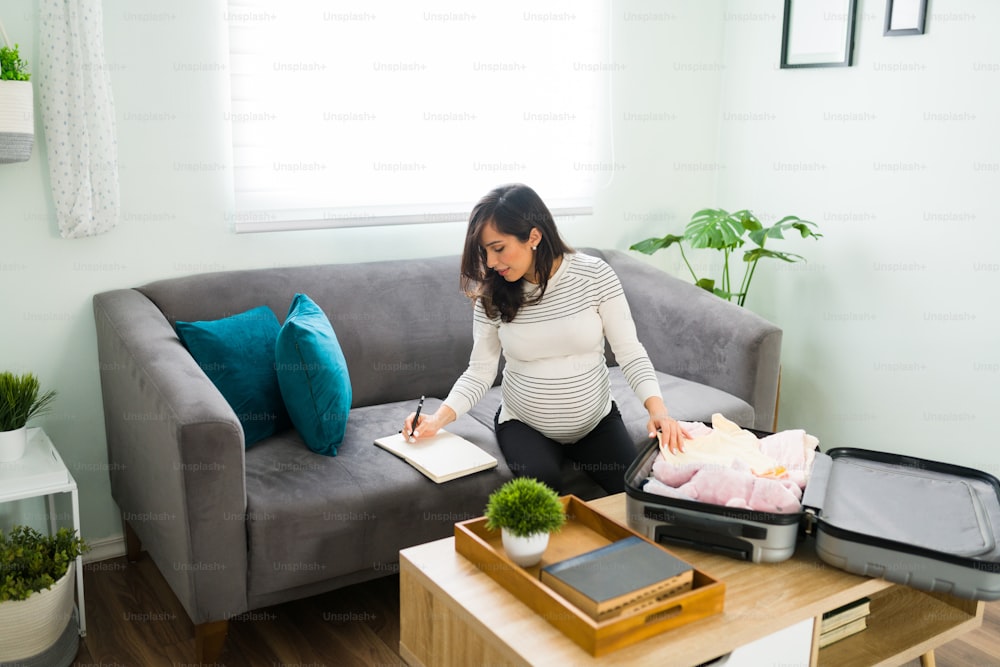 Excited pregnant woman in her 30s planning and writing on her agenda in the living room. Happy expectant mother preparing her hospital suitcase for the birth of her newborn