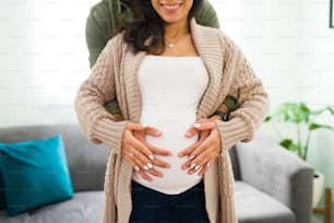 Stolzer hispanischer Mann, der eine glückliche schwangere Frau von hinten umarmt. Ehepaar, das mit seinem zukünftigen Baby den Bauch berührt