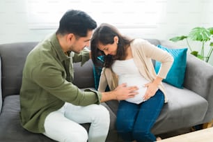 Une femme enceinte stressée a mal au ventre lorsqu’elle est assise sur le canapé. Mari alarmé essayant d’aider sa femme avec des contractions