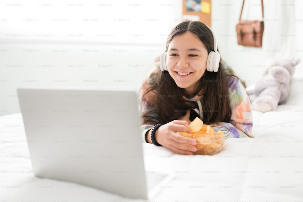 Happy teen girl with headphones watching a movie on her laptop while lying in bed. Smiling adolescent girl laughing and eating a bowl of potato chips