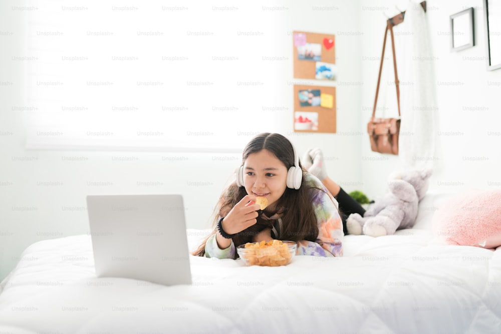 Linda adolescente menina desfrutando de um filme em seu laptop. Menina adolescente bonito assistindo vídeos on-line com fones de ouvido e comendo batatas fritas na cama
