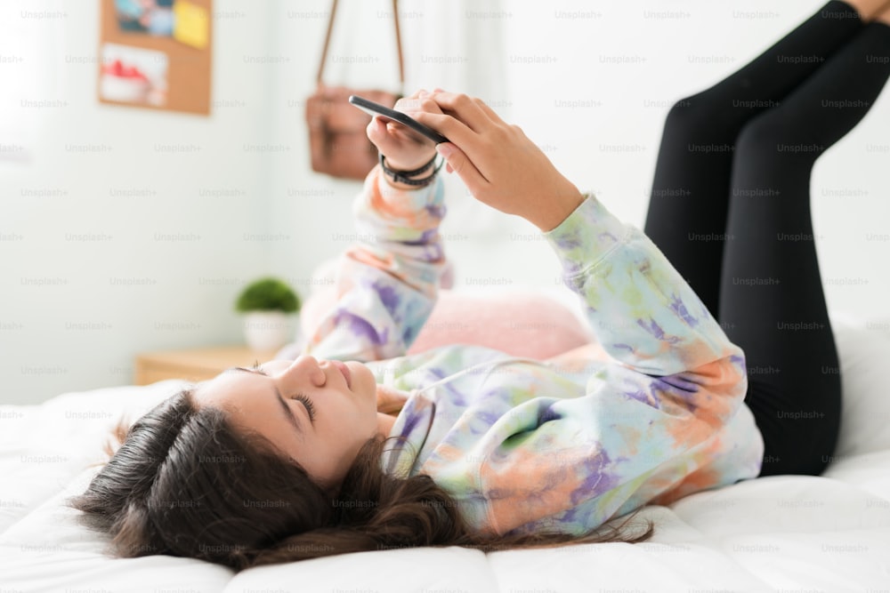 Good-looking adolescent girl lying in bed with her feet up in the wall. Caucasian teen girl scrolling through social media and texting on her smart phone