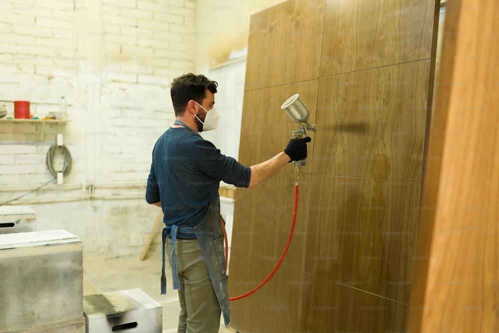 手袋とフェイスマスクを持つラテンの男性労働者がスプレーガンを持ち、木工所で茶色の木製のドアを塗る
