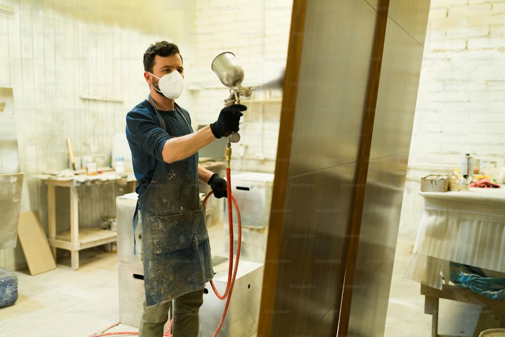 木工所の塗装ブースで働く30代のヒスパニック系男性。木製のドアにペンキをスプレーするフェイスマスクを持つハンサムな大工