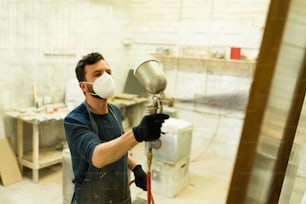 Uomo di bell'aspetto con una maschera facciale e guanti che usa una pistola a spruzzo per dipingere una porta di legno nella cabina di verniciatura di una falegnameria