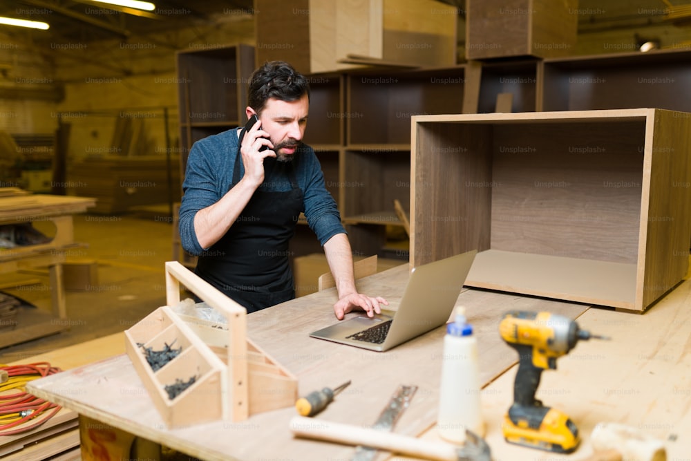 Good-looking male carpenter talking on the phone with a customer inside a woodshop. Hispanic man working on a carpentry project on his laptop