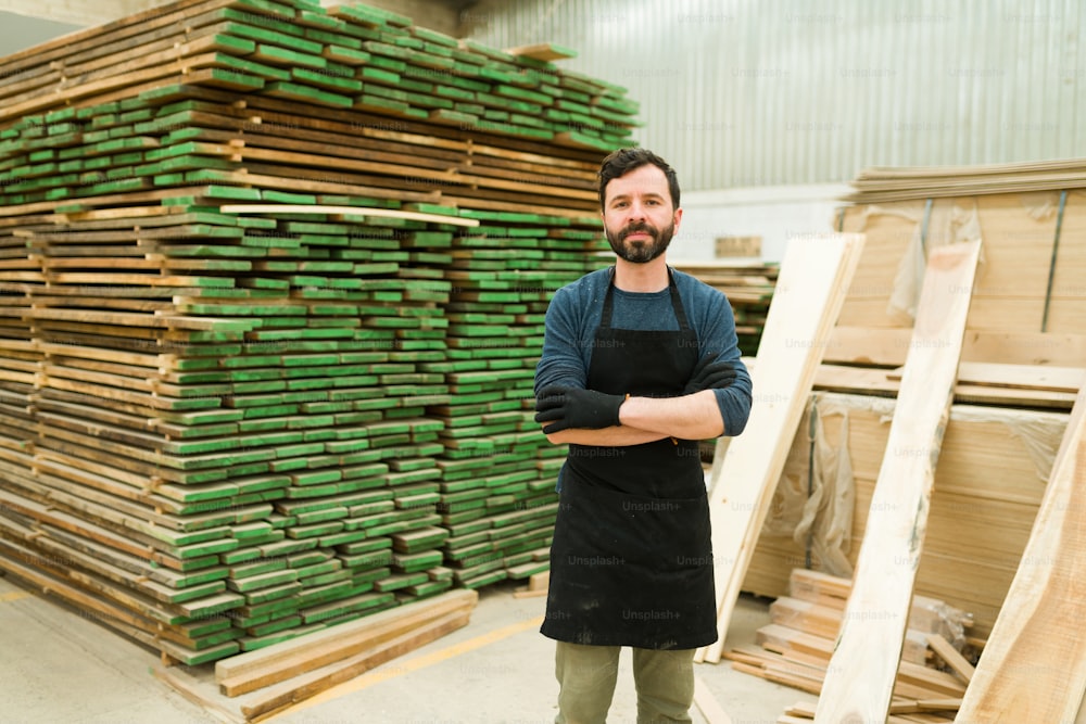 Attractive latin man in his 30s making eye contact with his arms crossed. Wood worker standing next to a pile of wooden bars inside a warehouse