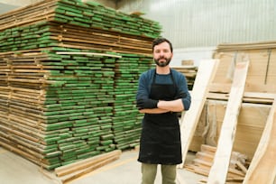 Attractive latin man in his 30s making eye contact with his arms crossed. Wood worker standing next to a pile of wooden bars inside a warehouse