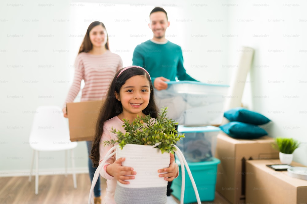 Beautiful elementary girl smiling and holding a green plant accompanied by her parents. Mom and dad holding boxes to unpack in their new home