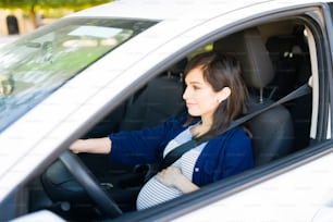 Seitenansicht einer hübschen schwangeren Frau, die ihr Auto mit angelegtem Sicherheitsgurt fährt. Frau in ihren 30ern lächelt und berührt ihren runden Bauch