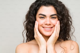 Mulher de boa aparência com cabelos encaracolados sorrindo e sentindo seu rosto limpo e fresco após sua rotina de cuidados com a pele