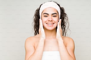 Happy beauty model wearing a towel and a headband and washing her face with a facial cleanser and soap
