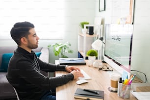 Side view of a male writer in his 20s sitting at his desk and typing on the computer. Hispanic man working and writing an online blog post