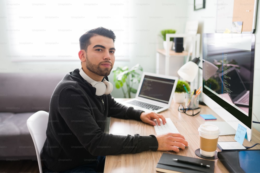 Handsome hispanic man in his 20s sitting on his desk at his home office. Young programmer working in software development and coding technologies