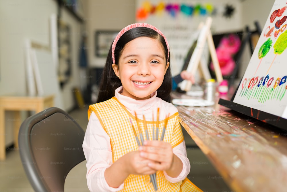 Beautiful hispanic elementary girl with an apron showing her paint brushes that she is using in her painting class in art school