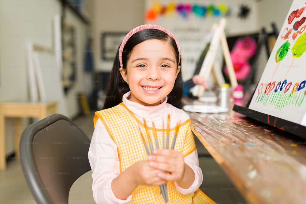 Bella ragazza elementare ispanica con un grembiule che mostra i suoi pennelli che sta usando nella sua classe di pittura nella scuola d'arte