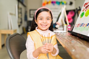 Belle fille hispanique du primaire avec un tablier montrant ses pinceaux qu’elle utilise dans son cours de peinture à l’école d’art