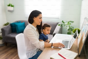 Pretty young single mom working at home office on a laptop while holding her baby boy