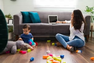 Giovane madre che parla al telefono e lavora su un computer portatile mentre il suo bambino gioca con i giocattoli in salotto