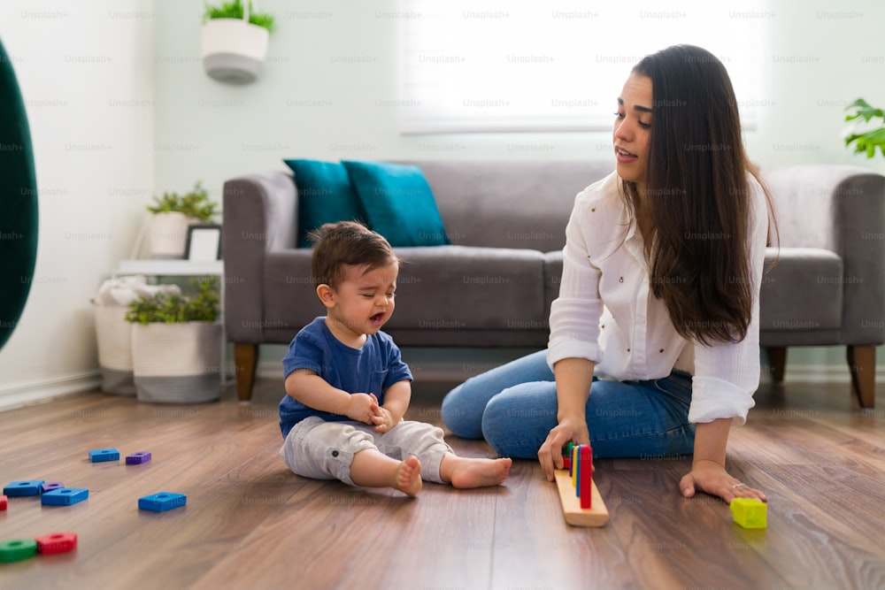 Baby boy sitting on floor and crying while playing toys with mother in living room