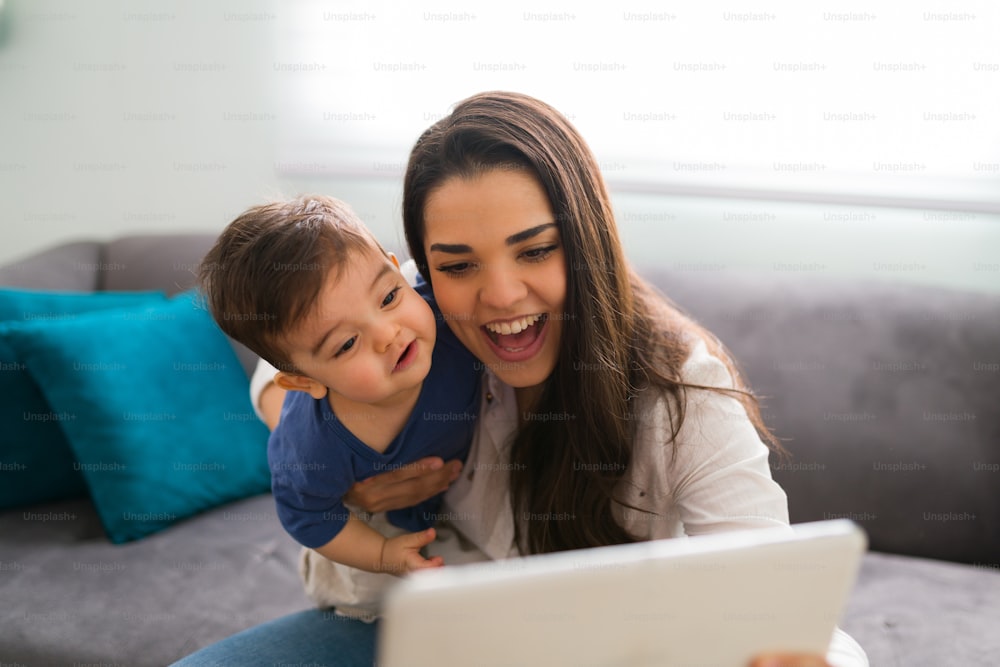 Beautiful young caucasian woman and her little son sitting at home looking at digital tablet and smiling