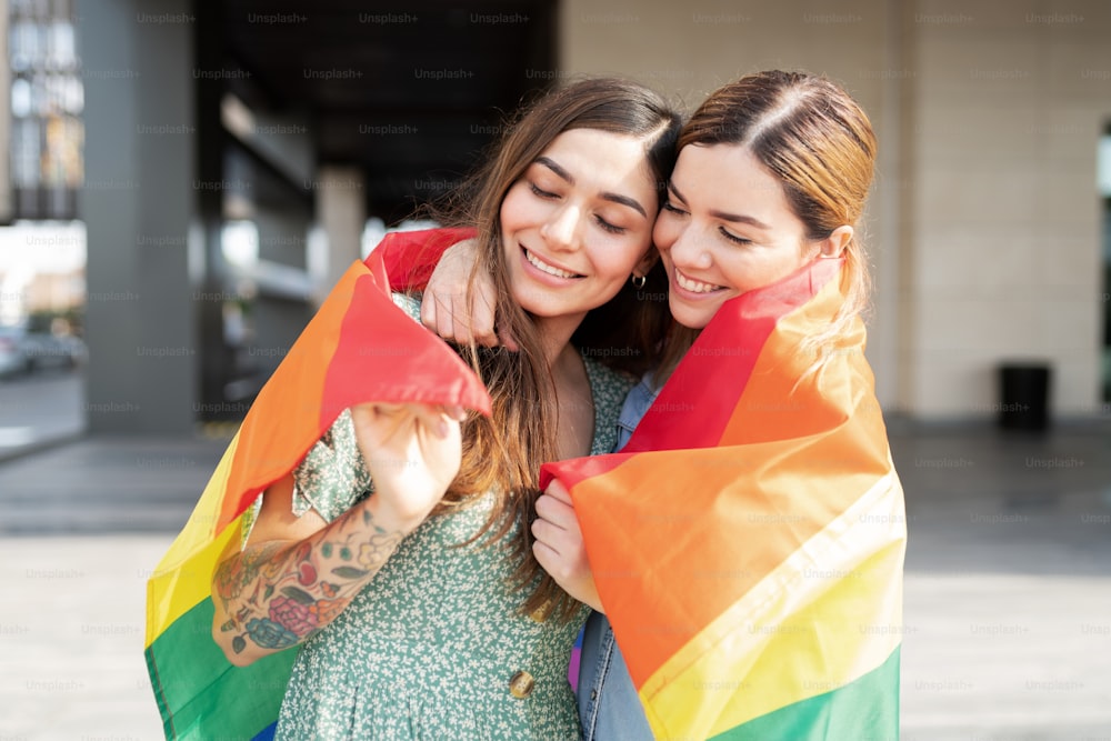 Happy gay couple cuddling with a rainbow flag around their shoulders and celebrating gay pride
