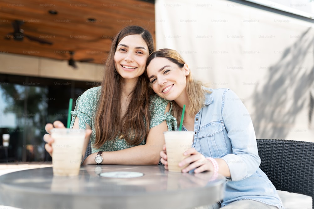 Caucasian young woman smimling and resting her head in the shoulder of her latin friend while drinking coffee