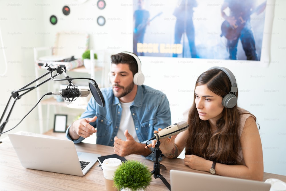 Attraktives Paar von Co-Moderatoren und Bloggern, die Kopfhörer tragen und mit ihren Mikrofonen sprechen, während sie einen Live-Podcast in einem Studio aufnehmen