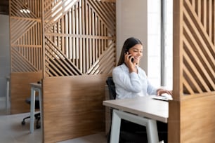 Happy young hispanic businesswoman talking over call phone while working on laptop at her workdesk