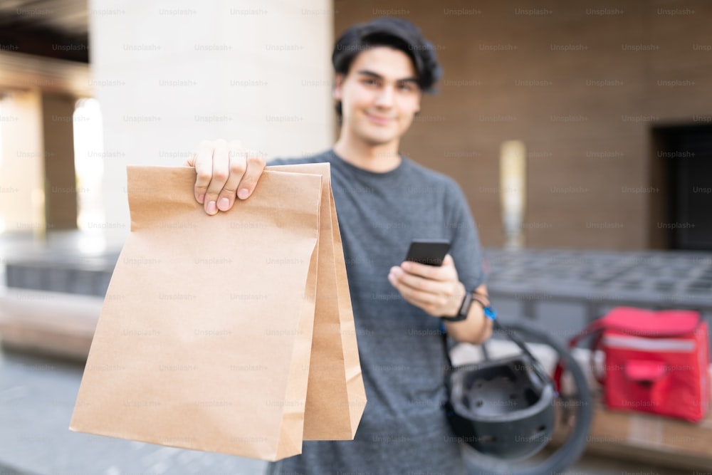 Hispanic young delivery man with mobile phone giving parcel in paper bags