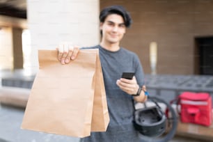 Joven repartidor hispano con teléfono móvil entregando paquete en bolsas de papel