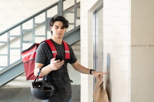 Latin delivery man using smartphone while pressing elevator button in building