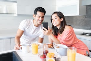 Casal jovem feliz olhando para o smartphone enquanto toma café da manhã em casa