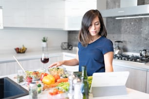 Mulher nova assistindo a receita on-line no tablet digital e preparando alimentos na cozinha