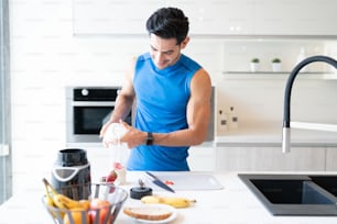 Young man pouring milk on strawberry slices in container at kitchen island before exercising