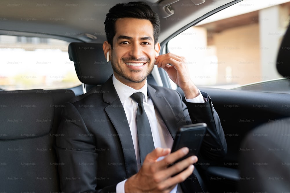 Hombre de negocios guapo sonriente escuchando música a través de auriculares inalámbricos mientras viaja en automóvil