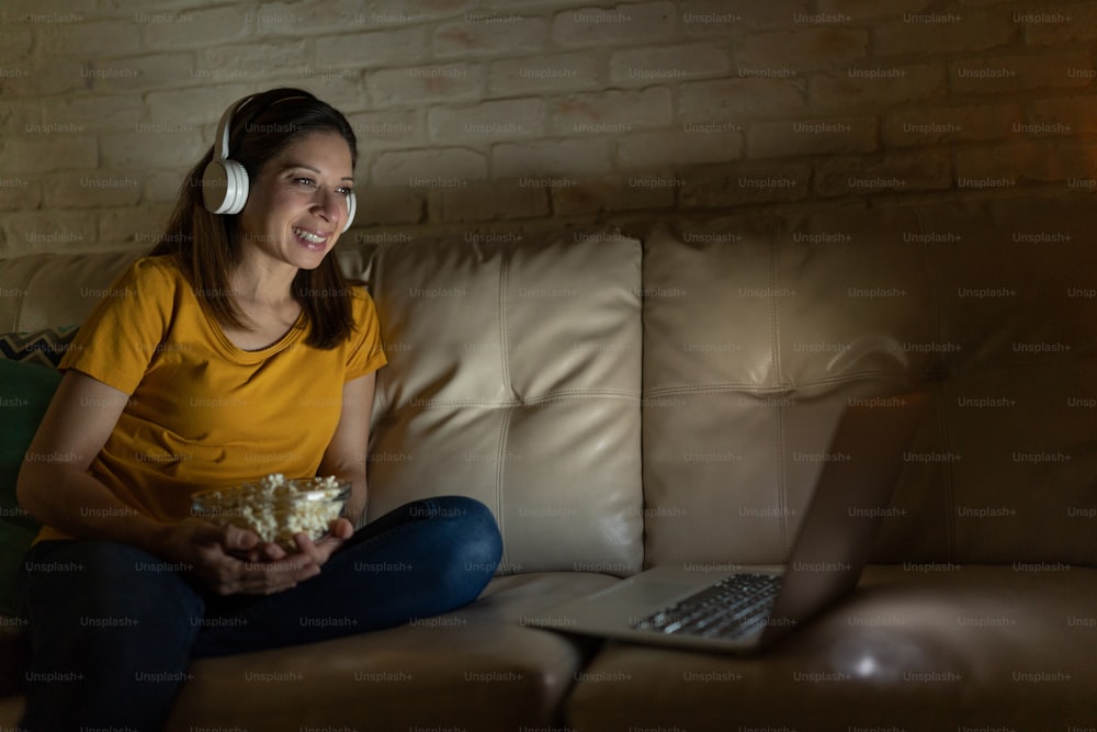 Retrato de uma mulher bonita que desfruta da noite de cinema em casa usando fones de ouvido e um laptop e sorrindo