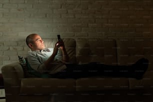 Vista di profilo di un uomo caucasico sdraiato su un divano di casa e che guarda il suo smartphone di notte
