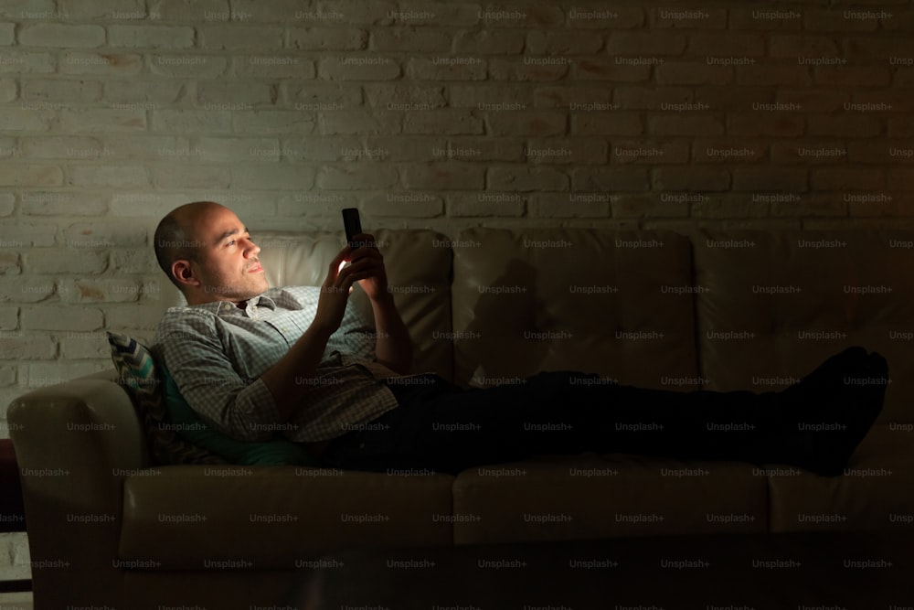 Profile view of a Caucasian man lying on a couch at home and looking at his smartphone at night