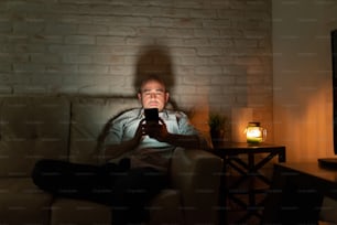 Lonely man in his 30s sitting on a couch at home and looking at his smartphone at night