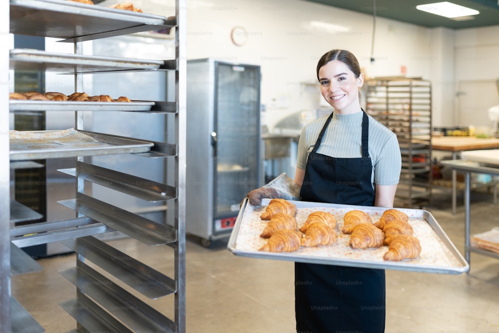Linda panadera caucásica sosteniendo una bandeja de pan recién horneado en una panadería y sonriendo