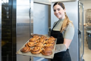 Hübsche junge Bäckerin, die Ofenhandschuhe trägt und ein Tablett mit frisch gebackenen Zimtschnecken in einer Bäckerei hält