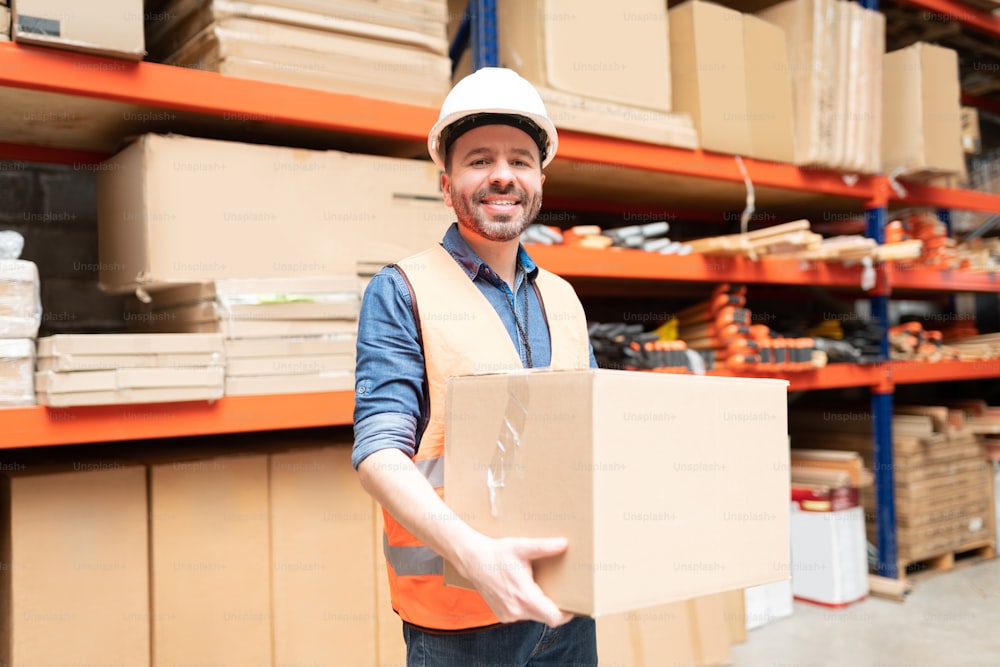 Happy good looking worker holding cardboard box while making eye contact against rack at factory