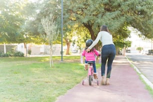 Rear View Of Mother Guiding Daughter In Riding Bicycle On Sidewalk At Park