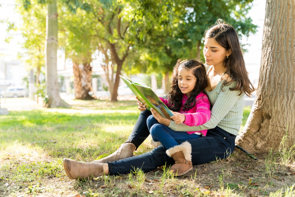 Junge Mutter liest ihrer Tochter ein Bilderbuch vor, während sie im Park gegen einen Baum sitzt