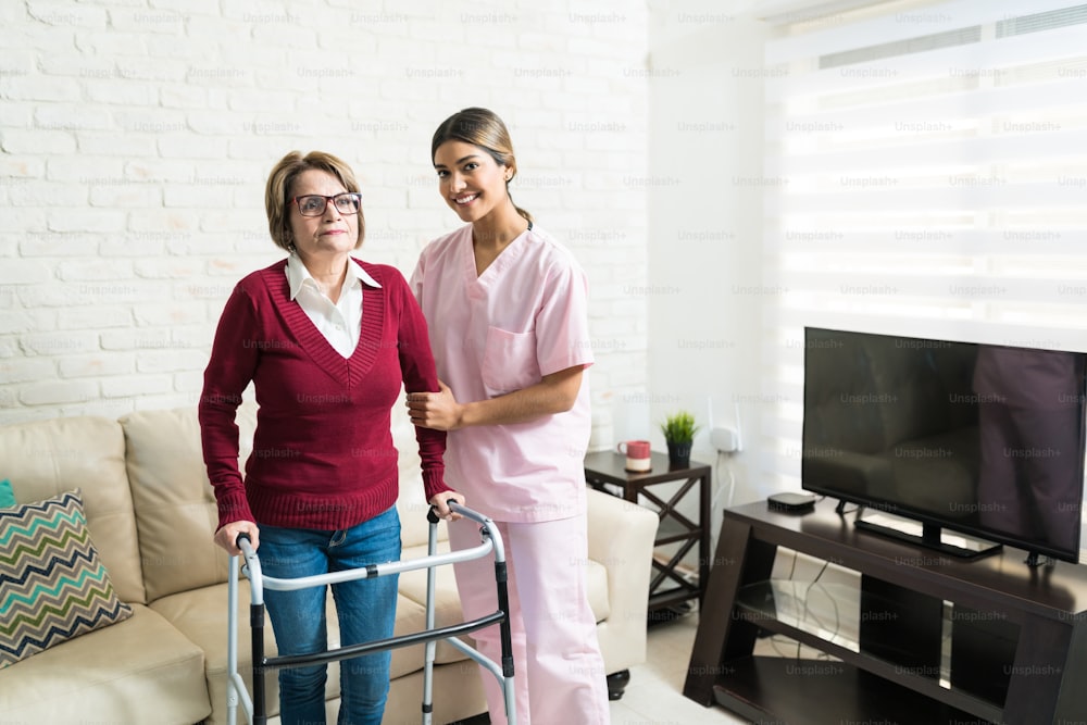 Smiling female caregiver helping elderly woman to walk with mobility walker at home